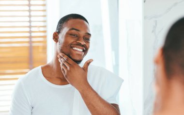 Male Skincare. Handsome Black Guy Looking In Mirror In Bathroom And Touching Beard, Attractive African American Man In White T-Shirt Enjoying His Appearance, Selective Focus On Reflection