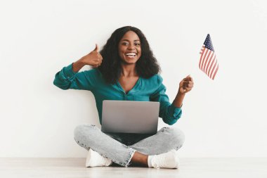 Pozitif genç siyah bayan, modern dizüstü bilgisayarı ve Amerikan bayrağıyla yerde oturuyor, kameraya başparmağını kaldırıyor ve gülümsüyor, online eğitim kursu öneriyor, beyaz stüdyo arka planı, kopyalama alanı.