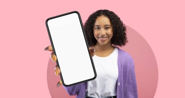 Genç bir kadın pembe bir arka plana karşı duruyor ve boş ekranlı bir akıllı telefonu sunmaktan mutluluk duyuyor. Beyaz bir gömlek ve lavanta rengi hırka giyiyor, pozitiflik yayıyor..