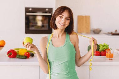 Diyet konsepti. Boynunda yeşil elma tutan ve kameraya gülümseyen, mutfaktaki iç kısımları kopyalayan, boynundaki şeritle mutlu formda kadın.