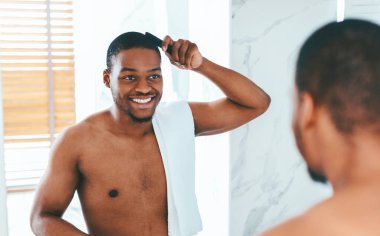 Yakışıklı, siyah bir adam banyoda saçlarını tarıyor ve aynaya bakıyor, mutlu bir Afrikalı Amerikalı adam sabahları hazırlanıyor, görünüşünü yansıtıyor ve eğleniyor, seçici bir şekilde odaklanıyor.