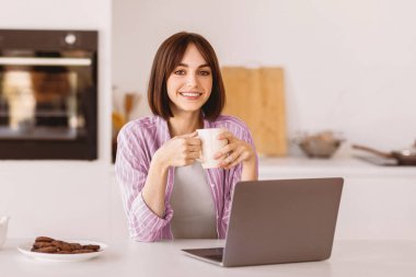 Laptop ve bardak kullanan mutlu bir kadının portresi, sabahları sıcak kahve içen, modern mutfak içinde oturan. Kadın video, film ya da webinar izliyor, kahvaltı yapıyor.