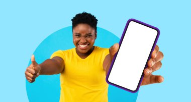 Neşeli bir birey, parmaklarını kaldırırken boş ekranlı bir akıllı telefonu tutuyor. Parlak mavi arka plan neşeli havayı güçlendirir, heyecan ya da kutlama hissi verir..