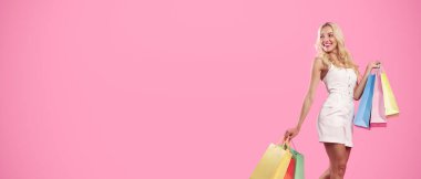 Büyük Satış ve İndirim. Moda kıyafetli mutlu kadın portresi ve beyaz spor ayakkabılar alışveriş çantalarıyla yürüyen, pembe stüdyo arka planında izole edilmiş fotokopi alanına bakan, panorama