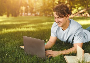 Diz üstü bilgisayar ve kitapla ödev yapan neşeli öğrenci dışarıda üniversite kampüsünde dinleniyor çimlerin üzerinde yatıyor. Eğitim ve teknoloji kavramı