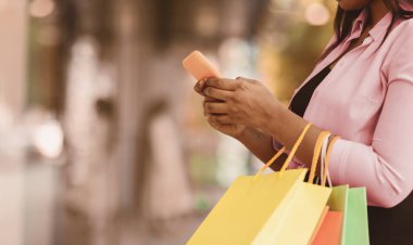 Tüketim ve Teknoloji. Tanımlanamayan siyah bir kadının akıllı telefon kullanması, alışveriş torbalarını tutması, dışarıda durması, kıyafet seçip sipariş etmesi, hediyeler alması, fotokopi alanı, bulanık arka plan.