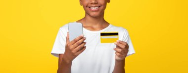 Beyaz tişörtlü genç siyah kız öğrencinin kredi kartı taşıması ve ödemeyi akıllı telefonla yapması, sarı arka planda izole edilmiş, boş bir alanda yapması güzel. Alışveriş koliği, mağazada online alışveriş, satın alma uygulaması