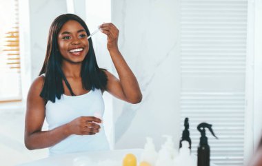 Skin Care Concept. Beautiful Black Female Applying Face Serum While Standing Near Mirror In Bathroom, Happy African American Woman Smiling At Her Reflection, Enjoying Domestic Beauty Treatments