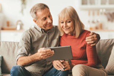 Senior Spouses Watching Movie Online On Tablet Computer Embracing Sitting On Sofa At Home. Happy Couple Browsing Internet Using Gadget Together. Retirement Leisure And Technology