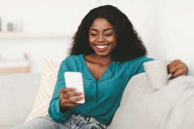 Rahat, çekici Afro-Amerikan bayan, güzel gür saçlı, modern cep telefonlu kanepe üzerinde dinlenen, kahve fincanı tutan, sosyal medyayı kontrol eden, gülümseyen, yakın plan, fotokopi çeken