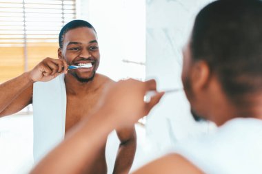 Diş bakımı. Banyoda dişlerini fırçalayan yakışıklı siyahi adam, evde sabah hijyeni sağlayan mutlu Afrikalı genç adam, yansımasına gülümseyen, seçici odaklı
