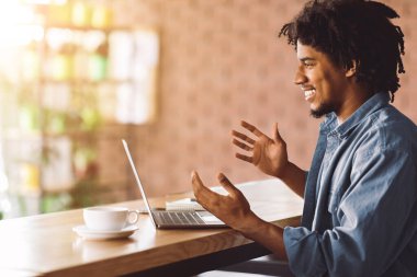 Gülümseyen duygusal kıvırcık siyah adam laptopta video çağrısı yapıyor, kafenin içindeki masada el kol hareketi ve iletişim kuruyor. Çevrimiçi toplantı, dijital göçebe, serbest çalışma, uzak ve modern teknoloji