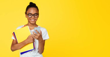 Gülümseyen Afro-Amerikan öğrenci gözlüklü ve gelişigüzel elinde kitaplarla akıllı telefondan, sarı arka planda izole edilmiş mesajlar tutuyor. Cihazla, modern eğitimle sonbaharda okula dönüyorum.