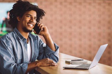 Gülümseyen, cep telefonuyla konuşan ve kafede dizüstü bilgisayarla çalışan Afro-Amerikan genç adam, keyifli mobil konuşmalar yapıyor ve bilgisayar kullanıyor, uzaktan kumandayla eğleniyor, boşluğu kopyalıyor.