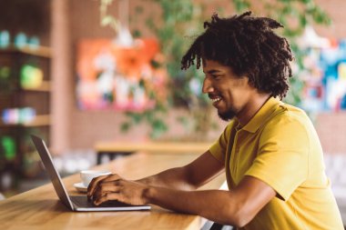 Gülümseyen kıvırcık, çekici siyah erkek bilgisayarda yazı yazıyor, kafenin içindeki sosyal ağlarda sohbet ediyor. Bağımsız yüklenici için uzak işyeri, serbest çalışma, modern iş ve teknoloji