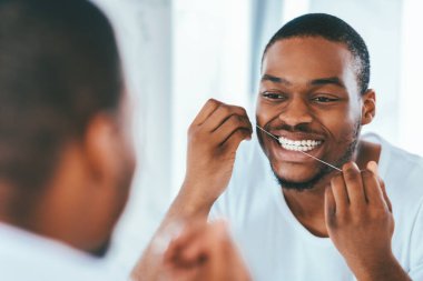 Ağız temizliği. Banyoda diş ipi kullanan Afrikalı genç adam, aynaya bakan ve dişlerini temizleyen siyah milenyum adamı, sabah hijyen rutini yapan, yansımaya odaklanan seçici bir bakış açısı.