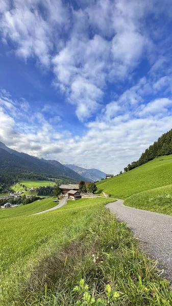 Santa Maddalena, Val di Funes, antika ev, dolambaçlı yol, yemyeşil otlaklar, kabarık bulutlu açık mavi gökyüzü, katmanlı Dolomitler-an Alp dinginliği tarafından çerçevelenmiş.