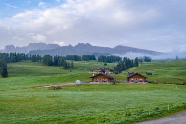 Sabah manzarası, yeşil çayırlarda dolambaçlı bir yol, büyüleyici kulübeler, ve Alpe di Siusi 'nin arka planı, Dolomitlerin kalbinde bir resim ve huzur sahnesi..