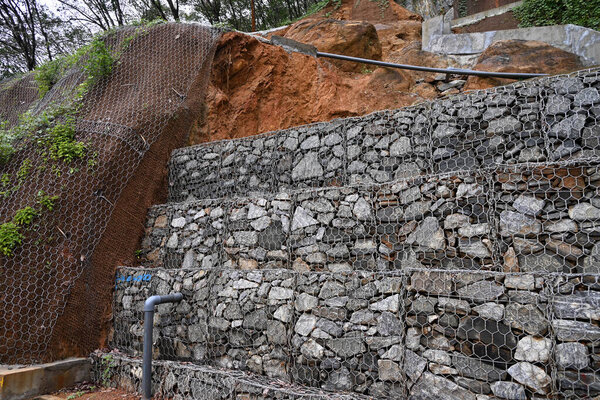 Вид на стеновые барьеры габиона, которые сделаны из шестиугольной сетки с гранитными валунами, чтобы предотвратить будущий оползень на тяжелом склоне, который недавно имел оползень