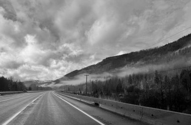 Etrafı sisli ormanlar ve tepelerle çevrili dolambaçlı bir dağ otoyolunun siyah beyaz fotoğrafı. Tepede dramatik bulutlar belirir, bu huzurlu, zamansız manzara gezmek, doğa ve minimalist temalar için kontrastı ve ruh halini yükseltir..