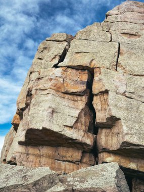 Yüzeyinde belirgin çatlaklar olan devasa bir kaya parçası, dağınık bulutlarla dolu canlı mavi bir gökyüzüne karşı uzanıyor. Kayanın sert dokusu ve toprak tonları doğal güzelliğini ve jeolojik önemini vurgular..