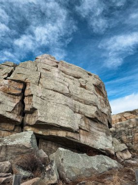 Keskin kenarları ve katmanlı dokuları olan devasa bir taş oluşumu ön plana hakim olur. Yumuşak ve dağınık bulutlarla dolu canlı bir gökyüzüne karşı kurulur. Kayalık detaylar ve açık gökyüzünün kontrastı çarpıcı bir doğal manzara yaratır..