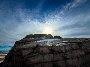 Okotoks Dengesizliğinin çarpıcı bir fotoğrafı. Alberta, Kanada 'da büyük bir buzul kayası. Görüntü, güneşin kayanın üzerinde yükseldiğini gösteriyor. Yumuşak, desenli bulutlarla süslenmiş canlı mavi gökyüzünün altında altın bir parıltı bırakıyor..