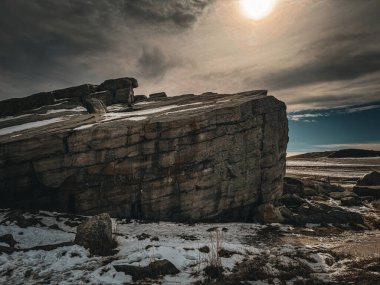Karlı bir manzarayla çevrili büyük, katmanlı bir kaya oluşumunun dramatik bir fotoğrafı kasvetli, bulutlu bir gökyüzü altında yumuşak güneş ışığıyla kırılıyor..