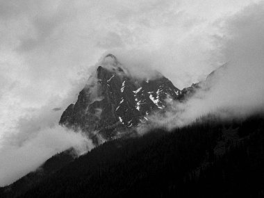 Yoğun sisin içinden çıkan keskin bir dağ zirvesi. Kayalık yamaçları karla kaplanmış. Düşük yamaçları kaplayan karanlık ormanlar, yukarıda dönen bulutlara dönüşüyor..