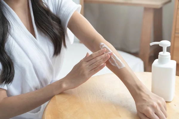 Young woman applying moisturizer cream to her arm in the morning routine.