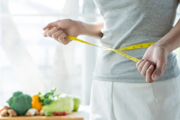 Eat good food for good shape concepts. Woman measuring her body by measure tape have a vegetables on the table as background. Girl checking her waist size down to follow up diet session result.
