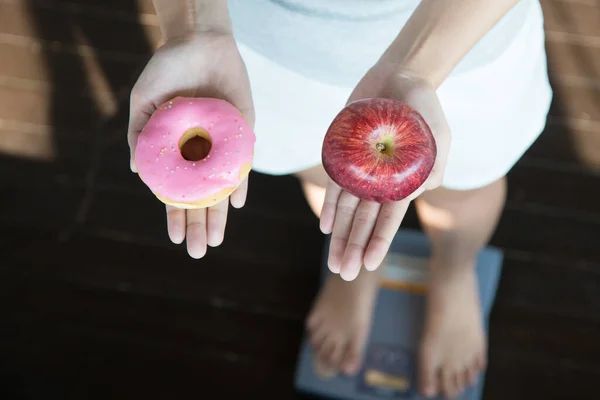 Choose a right choice for good health. Women is dieting comparison choice between donut and red apple during measuring weight on digital weight measure machine.