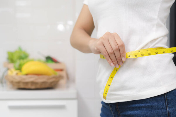 Eat good food for good shape concepts. Woman measuring her body by measure tape have a vegetables on the table as background. Girl checking her waist size down to follow up diet session result.