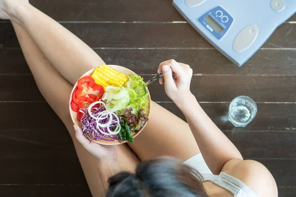 Woman dieting and control calories for good health concept. Above view of dieting woman sitting on the floor and eating homemade salad in the morning have a glass of water and weight scale beside her.