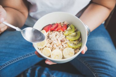 Eating healthy breakfast concept. Woman holding bowl of cereal and granola and variety fruits and glass of milk on th floor in the morning at home.