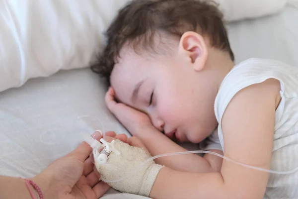 sick toddler in hospital holding mother's hand while sleeping