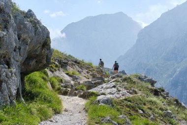 Sırt çantalı anonim erkek yürüyüşçülerin arka görüntüsü bulutlu mavi gökyüzüne karşı derin vadi ve yeşil çimlerle kayalık dağ formasyonunda yürürken 