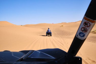 Arabanın arkasından, kasklı isimsiz bir adamın çölde ATV sürüşünü, güneşli bir günde Merzouga Çölü 'nde Morocco Afrika' da bulutsuz mavi gökyüzüne karşı kum tepeciklerini izledik.