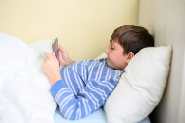 Boy lying in bed looking at a tablet, side view