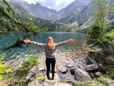 Polonyalı High Tatra Dağları 'ndaki Morskie Oko Gölü' nün önünde kolları açık duran beyaz, sarışın genç bir kadın kameraya bakmıyor.