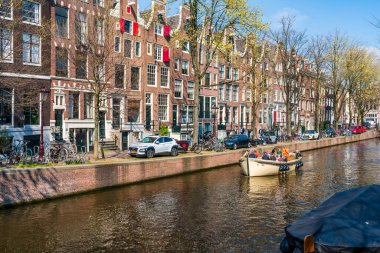 AMSTERDAM, HOLLLAND - 17 Nisan 2023: Turistler Amsterdam, Hollanda 'nın başkenti olan ve ayrıntılı kanal sistemi, dar evler ve bisiklet sistemiyle bilinen bir kanal boyunca tekne gezisinin keyfini çıkarıyorlar