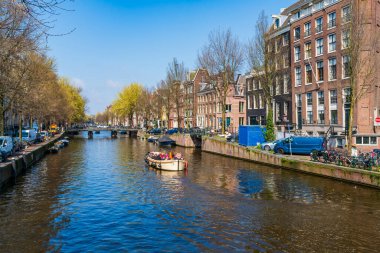 AMSTERDAM, HOLLLAND - 17 Nisan 2023: Turistler Amsterdam, Hollanda 'nın başkenti olan ve ayrıntılı kanal sistemi, dar evler ve bisiklet sistemiyle bilinen bir kanal boyunca tekne gezisinin keyfini çıkarıyorlar