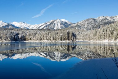 Kışın göldeki dağ yansıması. Karla kaplı ağaçlar ve mavi gökyüzü.