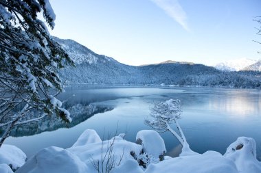 Kışın göldeki dağ yansıması. Karla kaplı ağaçlar ve mavi gökyüzü.
