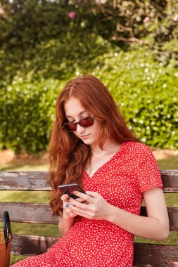 Odaklanmış kızıl saçlı, güneş gözlüğü takan bir kadın sıcak yaz günü dinlenirken bankta oturmuş akıllı telefonuyla sohbet ediyor.