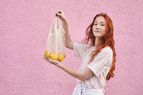 素食主义者快乐的女人拿着桔子水果包裹 同时在粉红的墙壁背景上摆出姿势 健康食品概念 — 图库照片