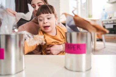 Down sendromlu bir çocuk evdeki masada annesinin yakınındaki çöpleri ayıklıyor. Geri dönüşüm konsepti için çöp toplama