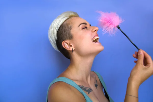 紫色の壁の背景の上に彼女の鼻にピンクの羽の棒で触れる間に積極的な入れ墨の女性は大声で笑っています エロ玩具のコンセプト ストックフォト — ストック写真
