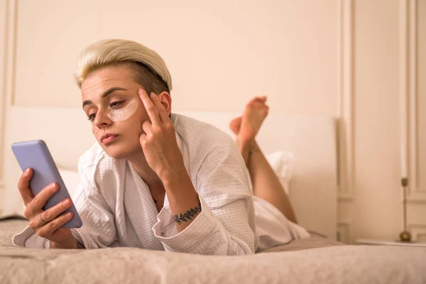 Серйозна Блондинка Читає Статтю Своєму Мобільному Телефоні Лежачи Біля Ліжка — стокове фото