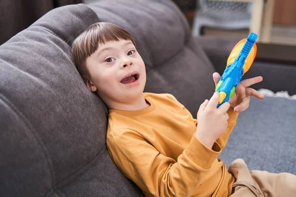 Portrait view of the overjoyed boy with genetic disorder sitting at the sofa and laughing while playing with toy gun. Happy childhood concept
